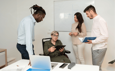 woman sitting with a tablet in hands and three employees standing around her listening