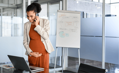 pregnant woman on phone holding her belly and looking at laptop with whiteboard behind her