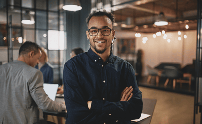 man with glasses smiling with arms crossed in office room with other people behind him