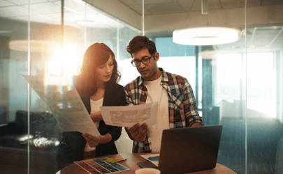 woman showing work documents to man on computer with the sunset showing through the window behind them