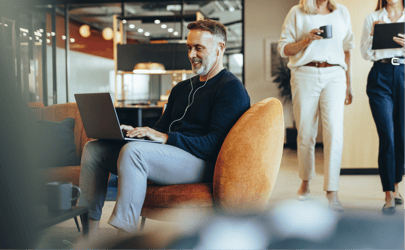 man sitting in office chair working on laptop with headphones while someone walks behind him