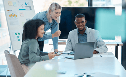 group of three business professionals working on a laptop together with a whiteboard behind them