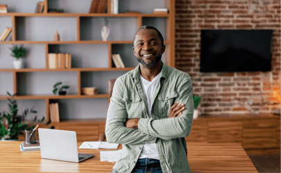 Man smiling in office with arms crossed and laptop behind him