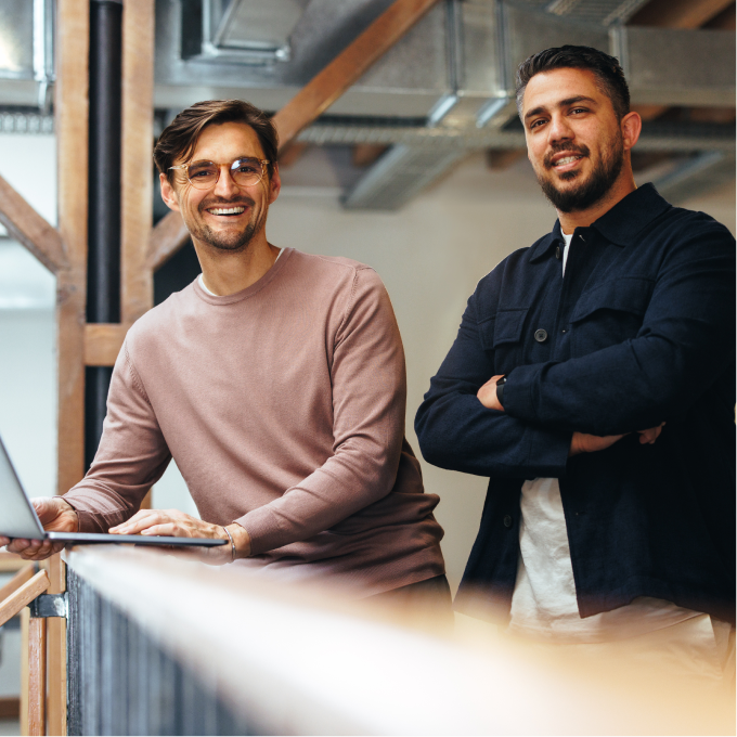 two men smiling and looking at camera in industrial building while one man holds laptop